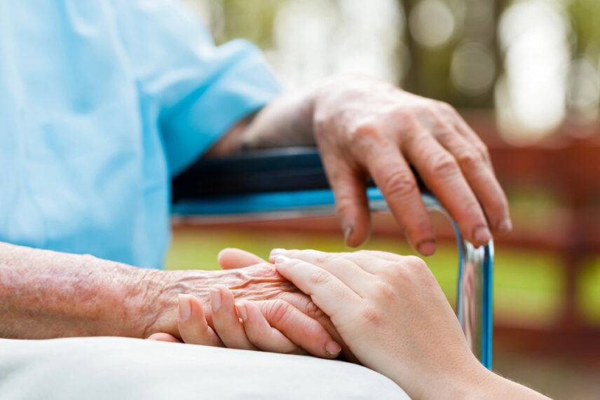 Caregiver holding elderly person’s hand in a supportive nursing home setting, representing long-term care planning and protection.